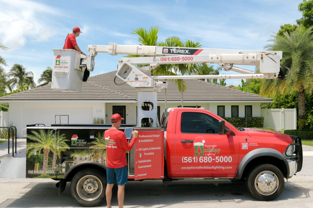 A big red bucket truck wrapped with imagery and information about the company called "Exceptional Holiday Lighting" and two workers, one on the floor and one on the bucket