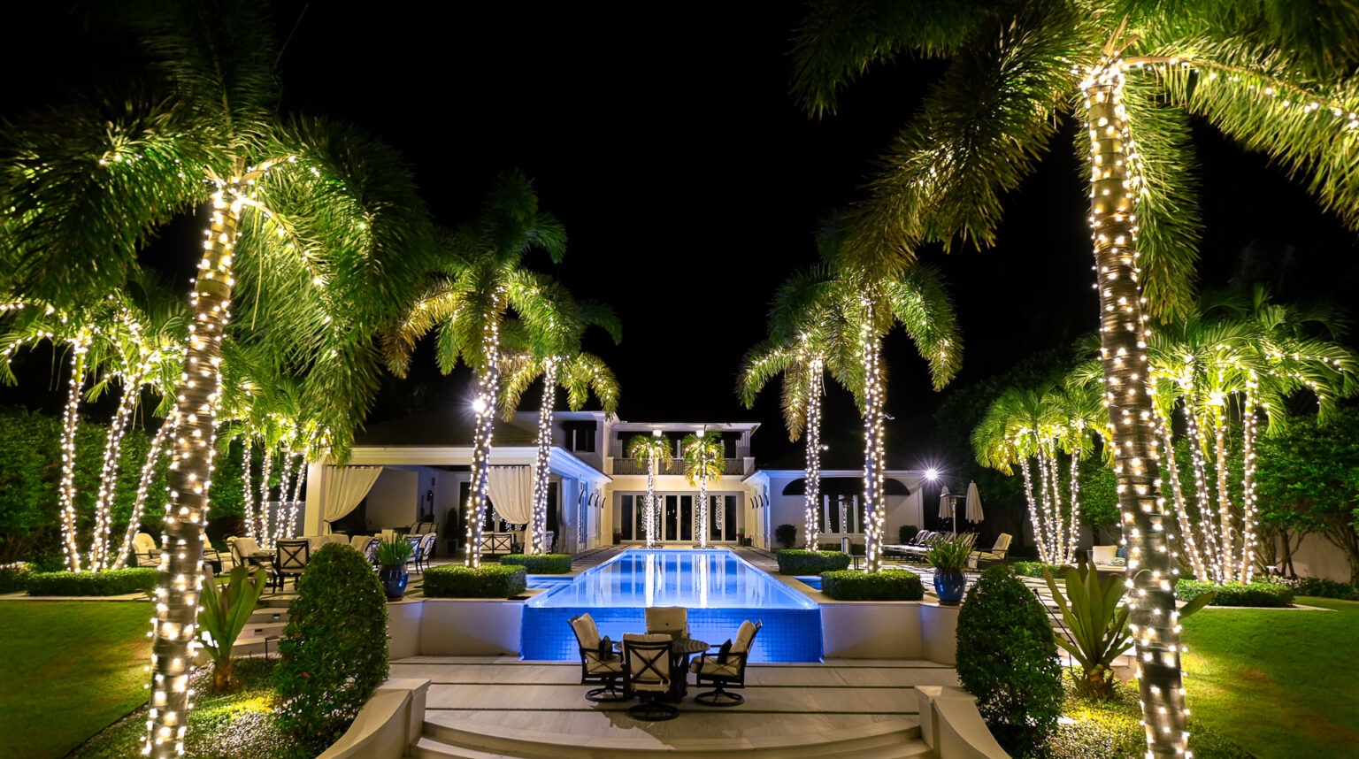 Stunning big modern house backyard with infinity pool in the middle and multiple palm trees running alongside the whole yard, decorated with the most professionally installed Holiday-style lighting in all of South Florida