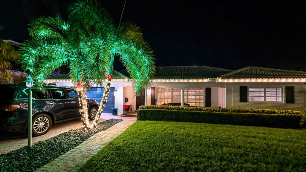 One-story house in Boca Raton Florida at nigh-time adorned in warm white holiday lights along the roofline and two small palm trees by the house entrance decorated with warm white, red, and green lights