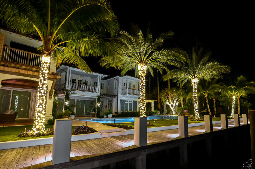 Luxurious holiday-style lighting installation on backyard palm trees of a high-end modern house with dock and water access in South Florida