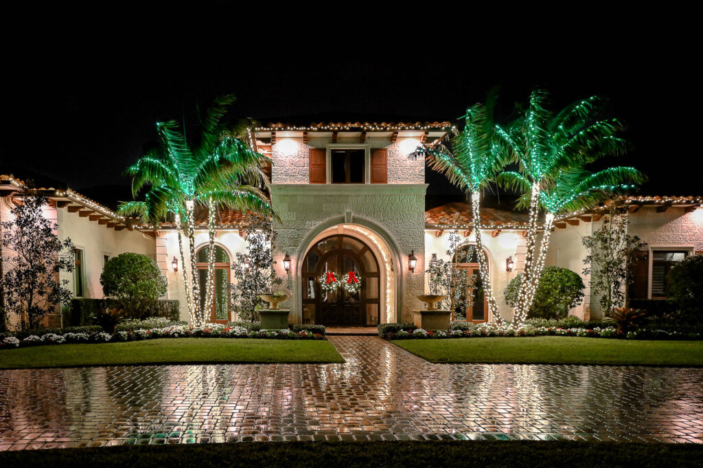 Holiday lighting installation on palm trees and entrance archway of a colonial-style house in Miami, Florida.