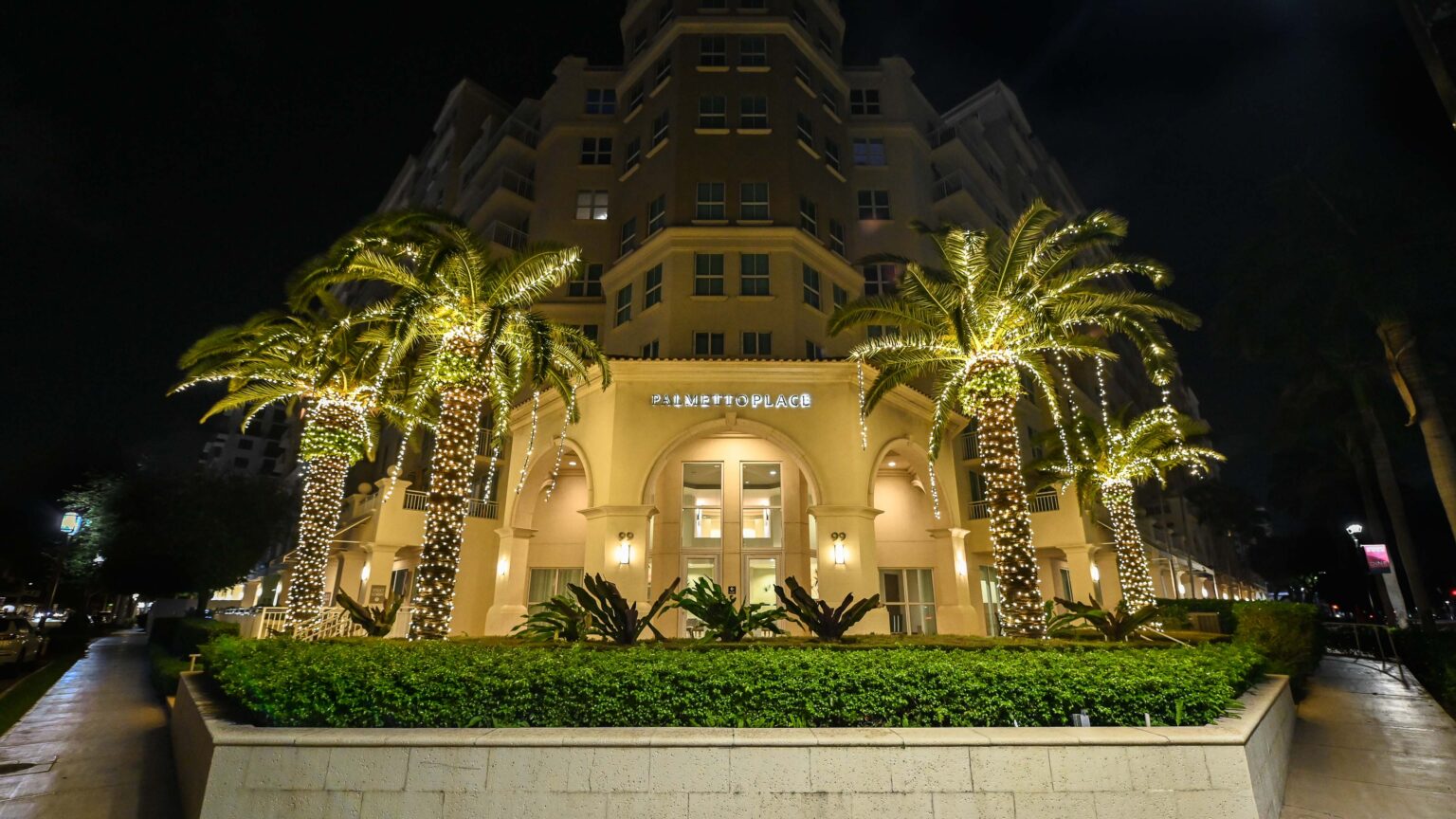 Front View of the entrance to the Palmetto Place building in Boca Raton, Florida. With four palm trees decorated with beautiful holiday lighting that illuminates the entrance and street.