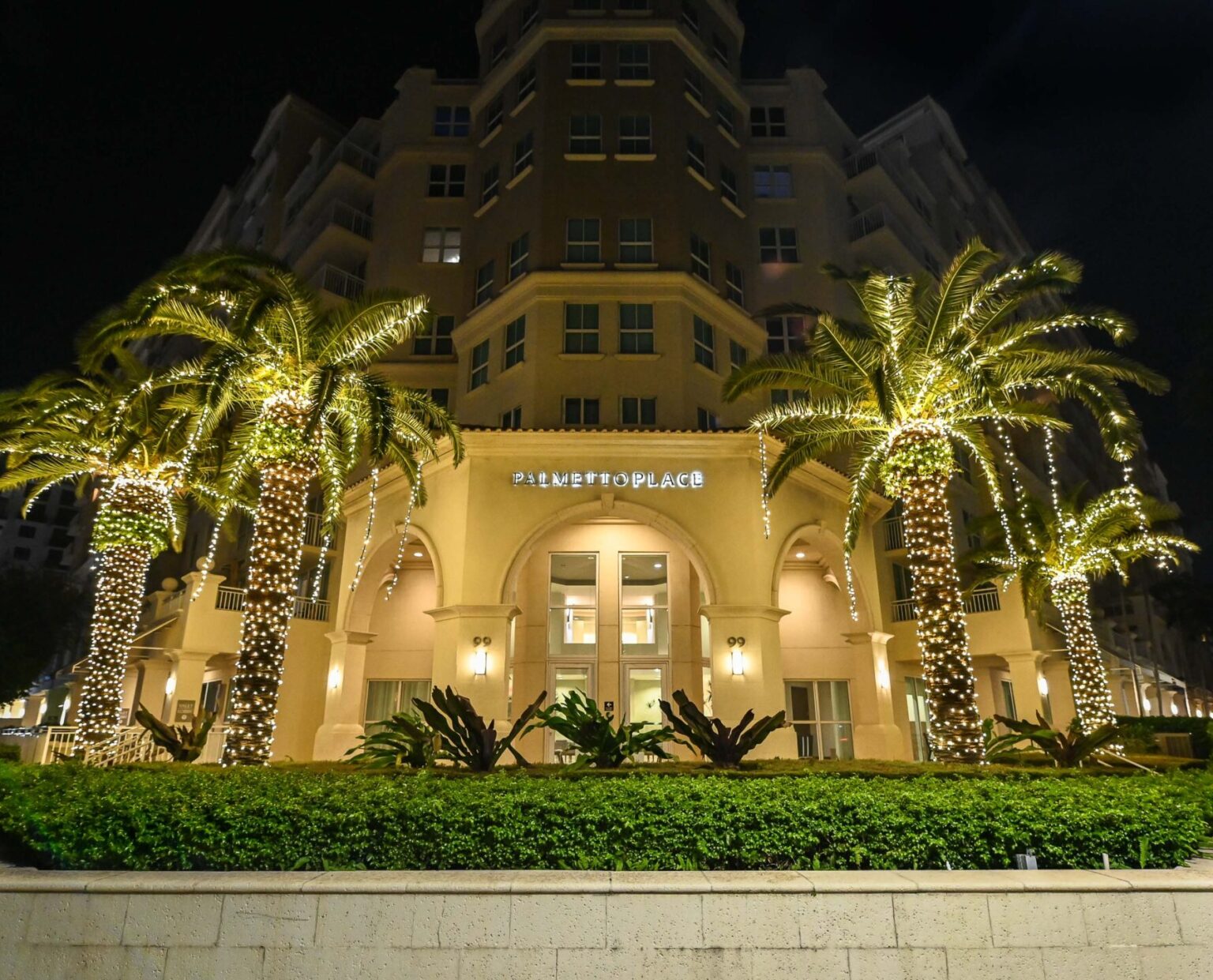 Front View of the entrance to the Palmetto Place building in Boca Raton, Florida. With four palm trees decorated with beautiful holiday lighting that illuminates the entrance and street.