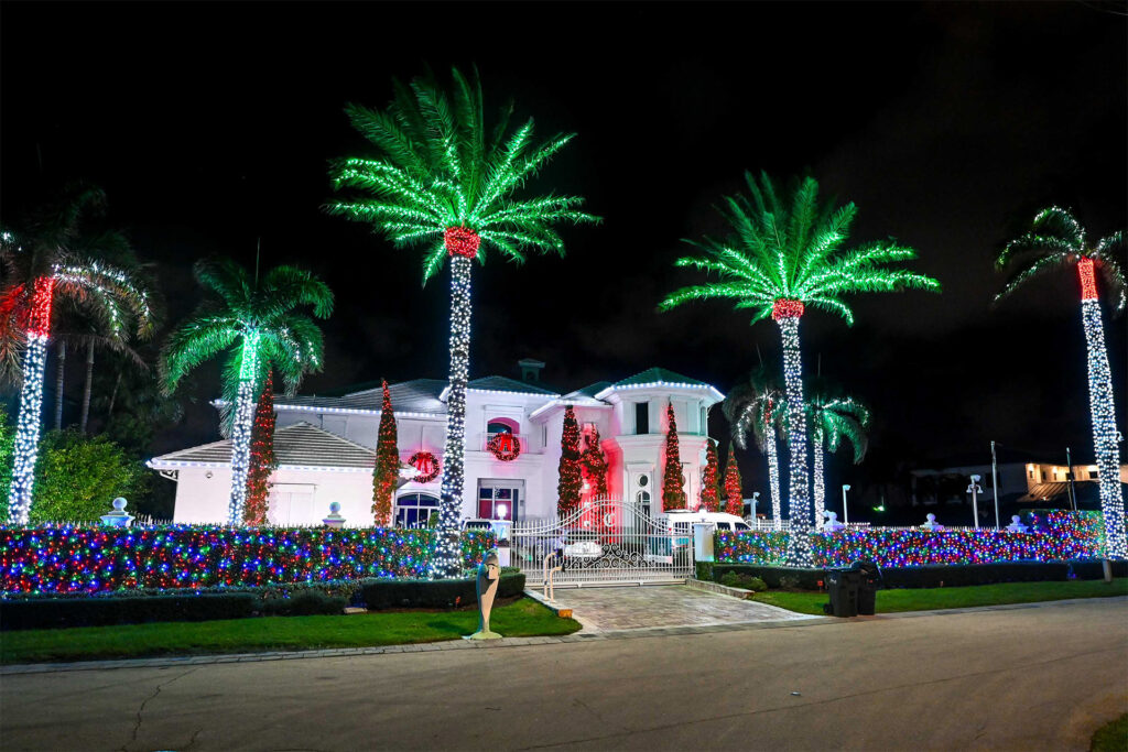 Breathtaking multi-color holiday light installation on a large luxury home with multiple palm trees, bushes, and wreaths decorated and lit up.