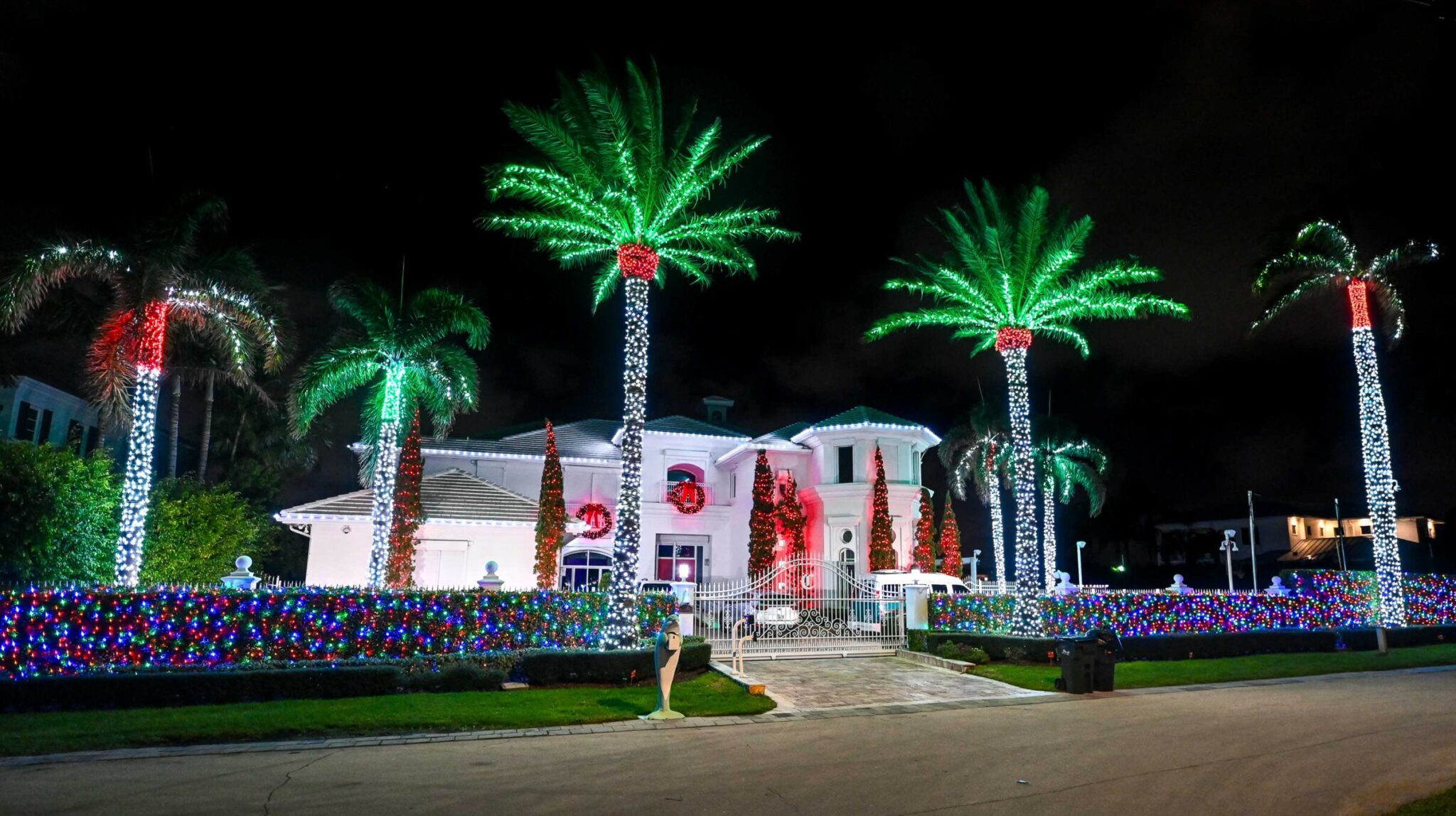 Breathtaking multi-color holiday light installation on a large luxury home with multiple palm trees, bushes, and wreaths decorated and lit up.