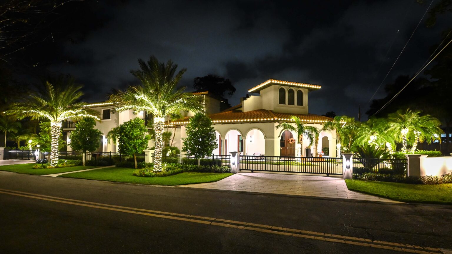 Big wealthy home decorated with warm white holiday lights around the roofline and many palm trees of different sizes all around the edges of the property