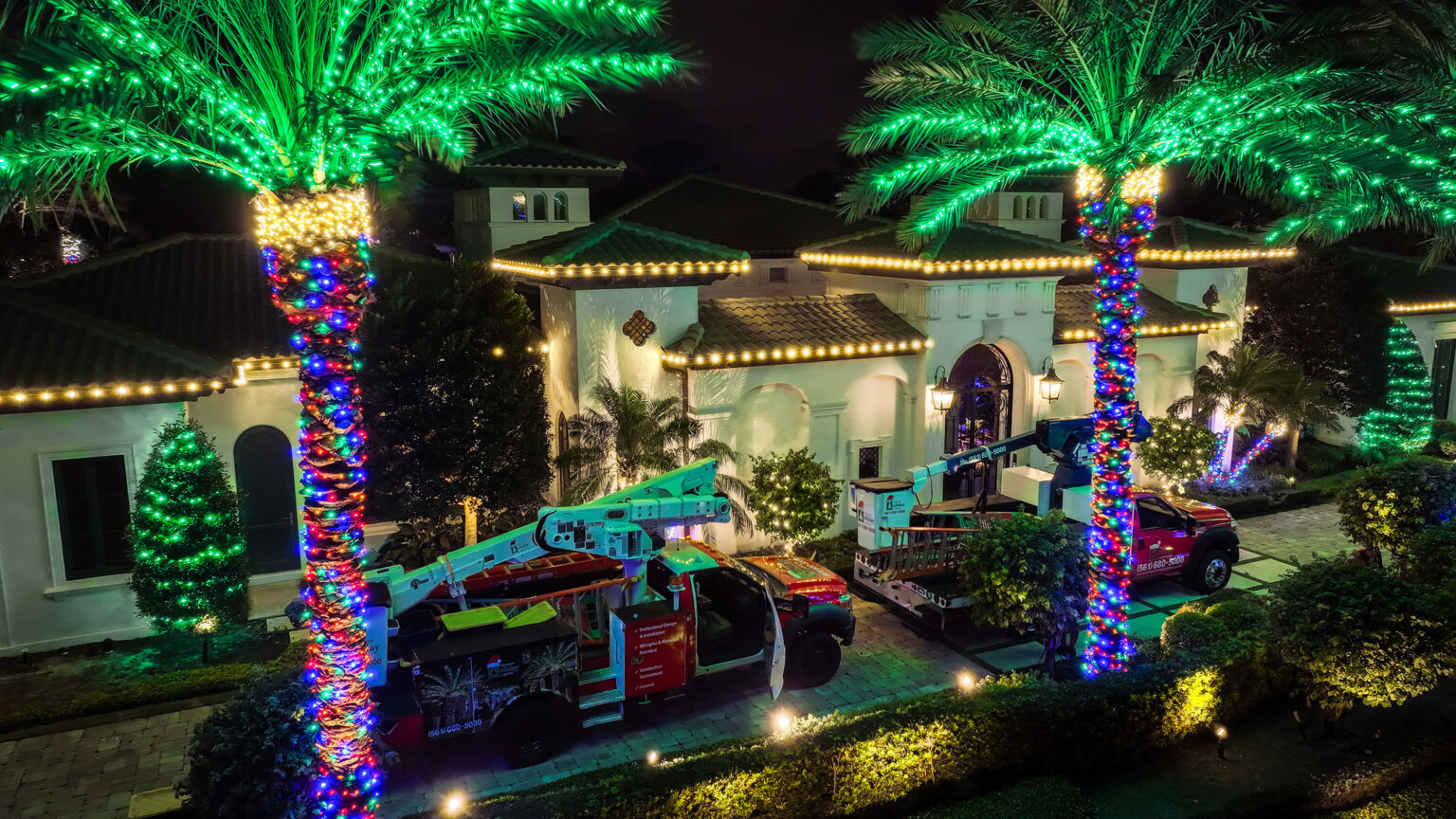 Breathtaking multi-color holiday light installation on a large luxury home with six palm trees decorated in front of the house, with lights wrapped around the roofline and bushes.