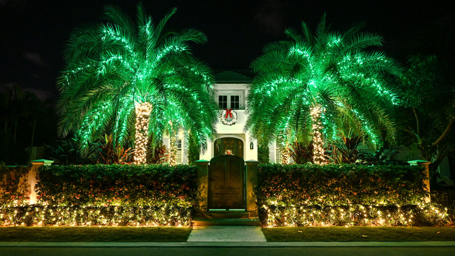 Symmetrically designed South Florida home with four palm trees in the front yard illuminated by the highest quality holiday-style lighting, with warm white trunks and lime green frond lights. Also, warm white holiday lights are decorating some of the bushes in the front area of the entryway facing the street, as well as an illuminated wreath on the house's central second-story balcony