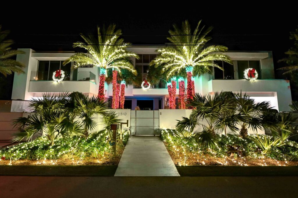 Big modern luxury home in Fort Lauderdale, Florida, with holiday-style lights installed on the symmetrically distributed 16 palm trees in the house's front yard, as well as the bushes and leaves in front of the house's street access entrance. Additionally, there are 3 symmetrically positioned Christmas-style wreaths lit up on the home's glass balconies.